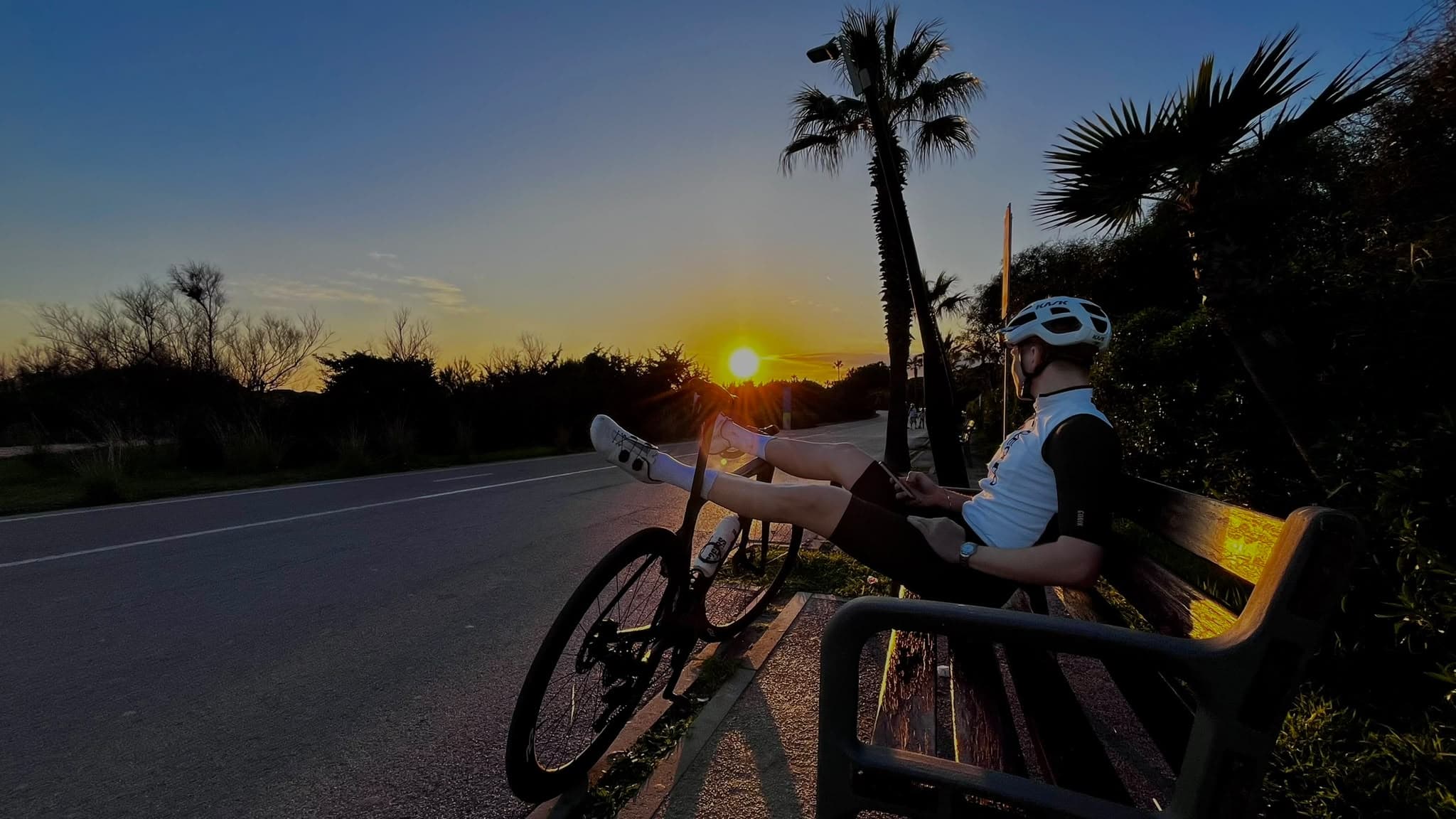 Un miembro del club descansando en un banco junto a su bici, con la puesta de sol de fondo en Castelldefels